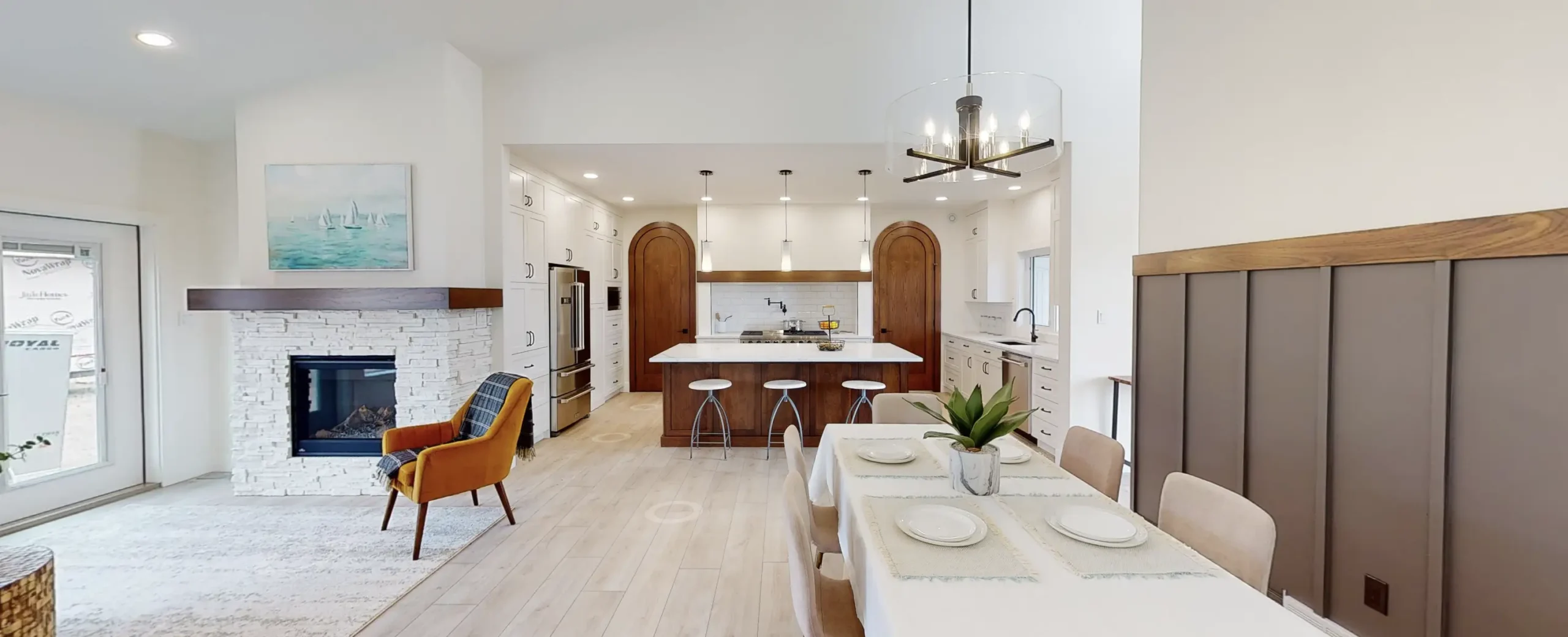 Open concept RTM home in Alberta or Saskatchewan with a white kitchen and arched wood doors, large island with three stools and pendant lights, white stone fireplace with wood mantel, mustard accent chair, and a dining table with modern chandelier.